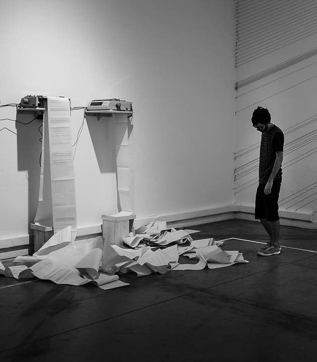 Black and white image of a man standing and looking down at a pile of printed papers spilling onto the floor from two wall mounted machines.