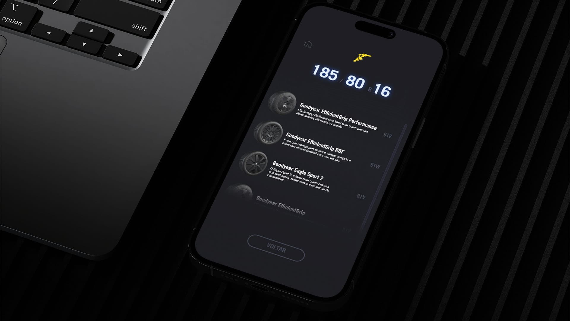 Um smartphone ao lado de um laptop sobre uma mesa. A tela do smartphone exibe a lista de produtos de pneus da Goodyear, mostrando títulos, descrições e detalhes de serviço para uma dimensão específica de pneu